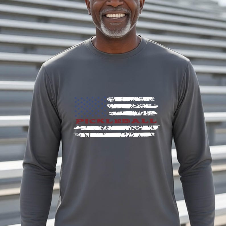 A charcoal gray long-sleeve shirt featuring a distressed American flag graphic in white and blue. The word PICKLEBALL is printed in red across the center of the flag design, which has a weathered, textured appearance on the fabric.