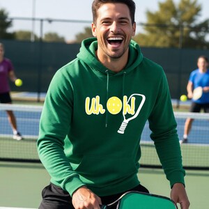 A forest green hooded sweatshirt featuring a white graphic of a pickleball paddle and a yellow pickleball with the text Uh oh! printed in white. The hoodie includes a front kangaroo pocket and matching green drawstring cords at the neck.