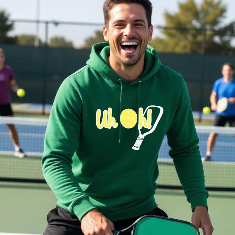 A forest green hooded sweatshirt featuring a white graphic of a pickleball paddle and a yellow pickleball with the text Uh oh! printed in white. The hoodie includes a front kangaroo pocket and matching green drawstring cords at the neck.