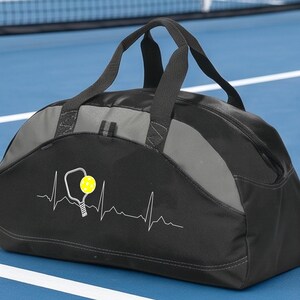 May include: A black and grey duffel bag featuring a white heartbeat line graphic with a pickleball paddle and a yellow pickleball. The bag has two black fabric handles and a zippered main compartment, constructed from durable synthetic material.