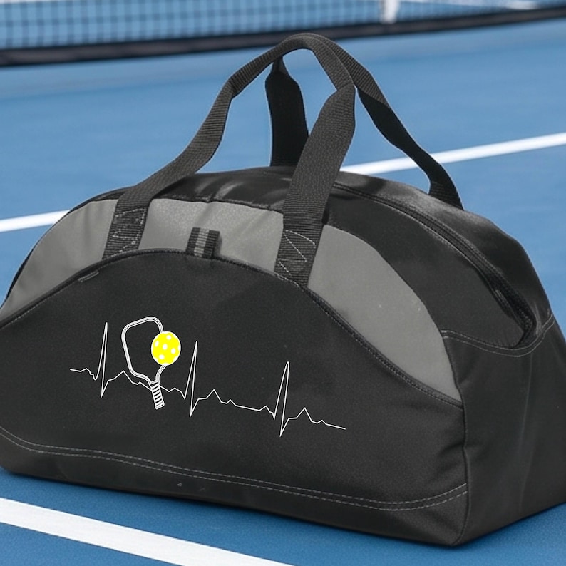 May include: A black and grey duffel bag featuring a white heartbeat line graphic with a pickleball paddle and a yellow pickleball. The bag has two black fabric handles and a zippered main compartment, constructed from durable synthetic material.