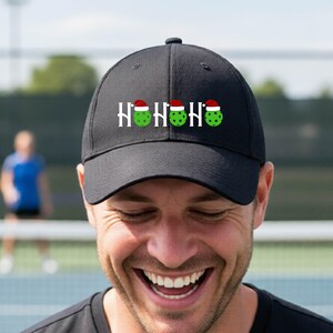 Könnte beinhalten: Eine schwarze Baseballkappe mit dem weißen Schriftzug HOHOHO, bei dem jeder Buchstabe O durch einen grünen Pickleball mit einer kleinen rot-weißen Weihnachtsmütze ersetzt ist. Die Kappe hat eine strukturierte Krone und einen gebogenen Schirm.