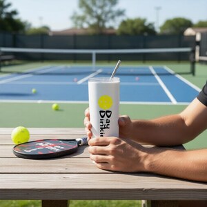 May include: A white cylindrical tumbler with a metal straw, featuring a yellow pickleball icon and the black text "Day Dinkin" printed vertically. The tumbler is held on a wooden surface next to a black pickleball paddle and a yellow ball.