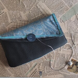 Può includere: Una pochette in tessuto blu e nero con un bottone decorativo e una catena argentata con un ciondolo. La pochette è in tessuto blu con un motivo bianco e una patta in pelle nera.