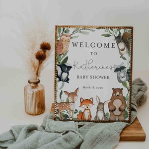 Może przedstawiać: Tablica powitalna na baby shower Katherine w stylu leśnym. Tablica ma białe tło z złotą ramką i zielony wieniec z r&oacute;żnymi zwierzętami leśnymi, w tym sową, szopem, jeleniem, lisem, kr&oacute;likiem i niedźwiedziem. Tekst na tablicy brzmi "Witamy na baby shower Katherine" i "18 marca 2020".