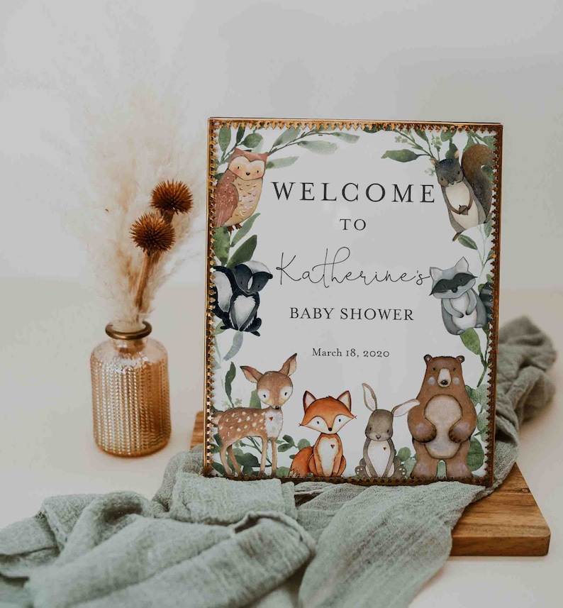 Może przedstawiać: Tablica powitalna na baby shower Katherine w stylu leśnym. Tablica ma białe tło z złotą ramką i zielony wieniec z r&oacute;żnymi zwierzętami leśnymi, w tym sową, szopem, jeleniem, lisem, kr&oacute;likiem i niedźwiedziem. Tekst na tablicy brzmi "Witamy na baby shower Katherine" i "18 marca 2020".