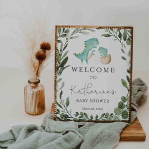 May include: A welcome sign for a baby shower with a green and gold frame. The sign features a watercolor illustration of two dinosaurs, one green and one blue, with the text "Welcome to Katherine's Baby Shower March 18, 2020".