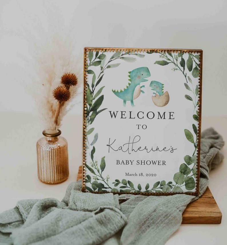 May include: A welcome sign for a baby shower with a green and gold frame. The sign features a watercolor illustration of two dinosaurs, one green and one blue, with the text "Welcome to Katherine's Baby Shower March 18, 2020".
