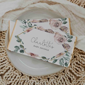 Pode incluir: Uma caixa de presente branca com uma moldura geométrica dourada e um design floral. A caixa tem o texto "Charlotte's Baby Shower" impresso.