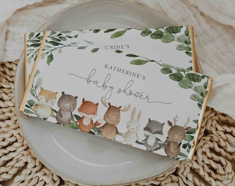 May include: A white chocolate bar wrapper with a green leafy design and a line of woodland animals. The text "Katherine's baby shower" is printed on the wrapper.