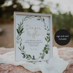 May include: A framed sign for a diaper raffle, featuring elegant gold script and a wreath of green leaves. The sign includes instructions and a thank you message. The sign is displayed on a textured surface with a soft, white fabric.