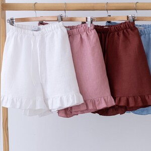 May include: A wooden clothing rack displays five pairs of linen shorts in various colors: white, pink, burgundy, light blue, and dark green. Each pair has a ruffled hem and an elastic waistband.