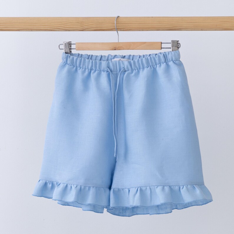 May include: Light blue linen shorts with a drawstring waist and ruffled hem, hanging on a wooden hanger. The shorts are made of a soft, lightweight fabric, perfect for warm weather. The shorts are a solid light blue color.