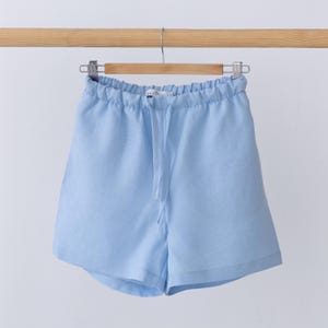 May include: Light blue shorts featuring an elasticated waistband with a matching drawstring tie. The shorts have a relaxed fit with a simple hemline and are displayed hanging from a wooden hanger against a plain, neutral background.