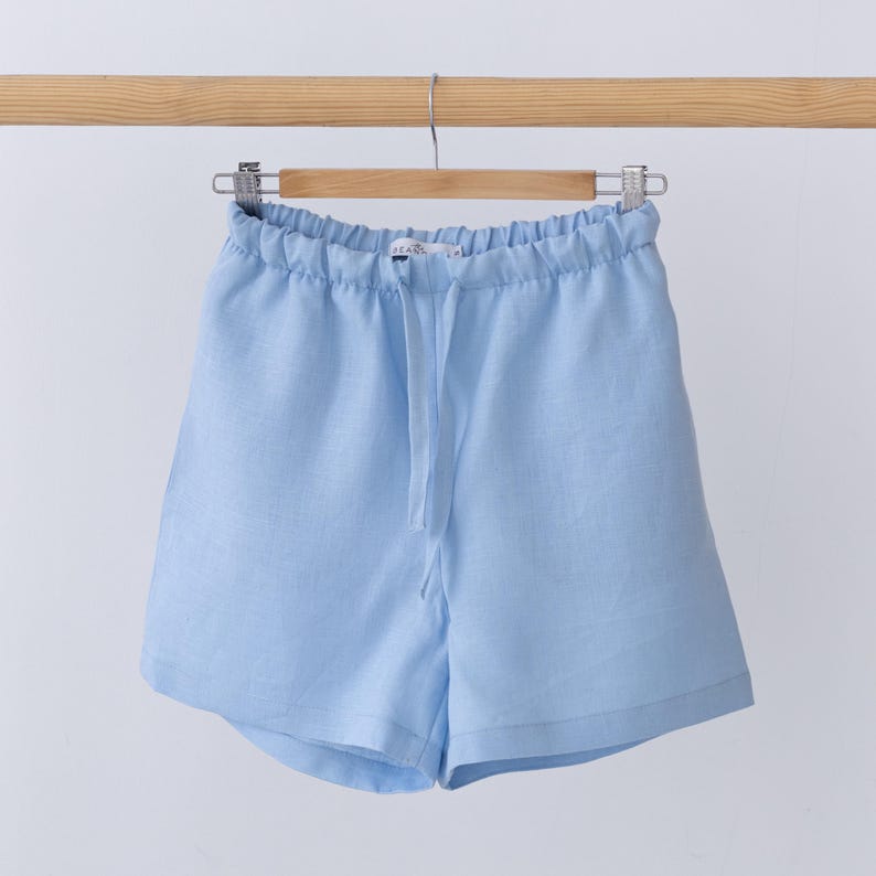 May include: Light blue shorts featuring an elasticated waistband with a matching drawstring tie. The shorts have a relaxed fit with a simple hemline and are displayed hanging from a wooden hanger against a plain, neutral background.