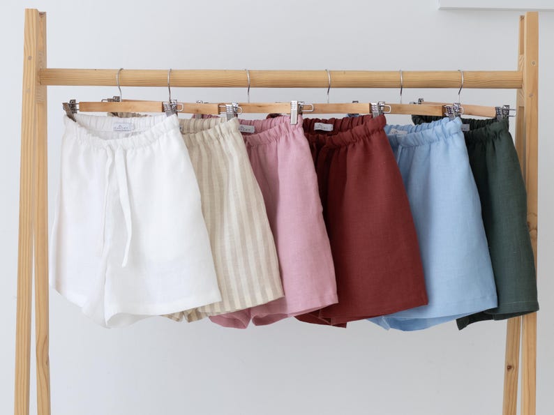 May include: A collection of six pairs of shorts hanging on a wooden rack. The shorts feature an elastic waistband with a drawstring and come in white, beige striped, light pink, terracotta, light blue, and dark green solid colours.
