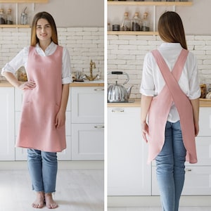 May include: A pink linen apron with a criss-cross back strap and two pockets. The apron is shown being worn by a person in a kitchen setting.