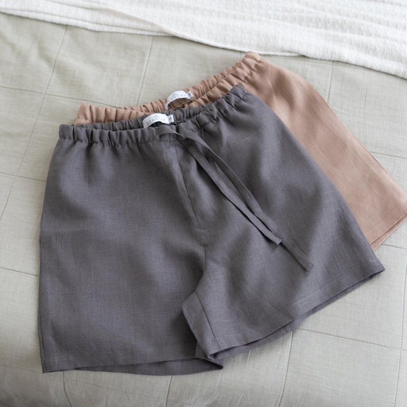 May include: Two pairs of pull-on shorts with elastic waistbands and drawstring ties. One pair is dark charcoal grey and the other is a light tan beige. Both shorts feature a visible white brand label sewn into the interior centre back waistband.