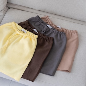 May include: A set of four pairs of shorts in yellow, dark brown, charcoal grey, and beige. Each pair features an elasticated waistband with a drawstring, a simple hem, and a small white interior label with the text ANCHY visible at the waist.