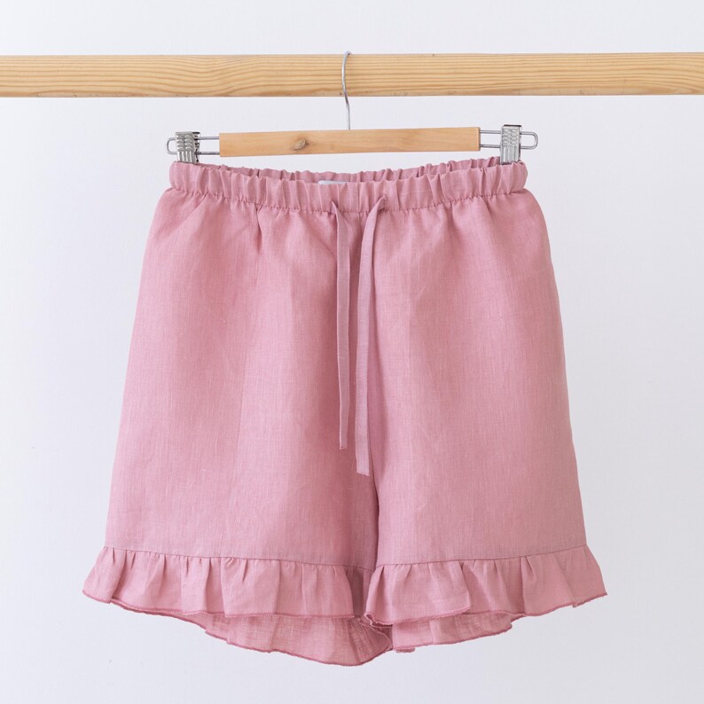 May include: Pink linen shorts with a drawstring waist and ruffled hem, hanging on a wooden hanger. The shorts are a solid color and appear to be made of a lightweight fabric, suitable for warm weather.