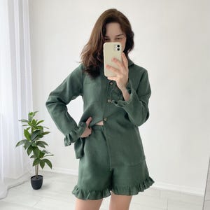 May include: A green linen pajama set featuring a long-sleeved button-down shirt and matching shorts with ruffled hems. The shirt has a collar and wooden buttons. The shorts have an elastic waistband. The set is made of a lightweight fabric.