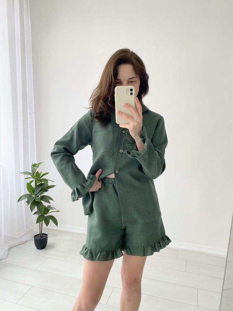 May include: A green linen pajama set featuring a long-sleeved button-down shirt and matching shorts with ruffled hems. The shirt has a collar and wooden buttons. The shorts have an elastic waistband. The set is made of a lightweight fabric.