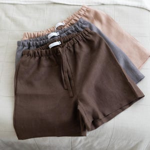 May include: Three pairs of shorts stacked on top of each other, featuring an elastic waistband with a drawstring. The shorts are coloured in dark brown, charcoal grey, and light beige, each with a white brand label sewn inside the back waist.