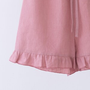 May include: Close-up of pink shorts with a ruffled hem. The shorts are made of a lightweight fabric and feature a drawstring waist. The shorts are a solid color and appear to be new.