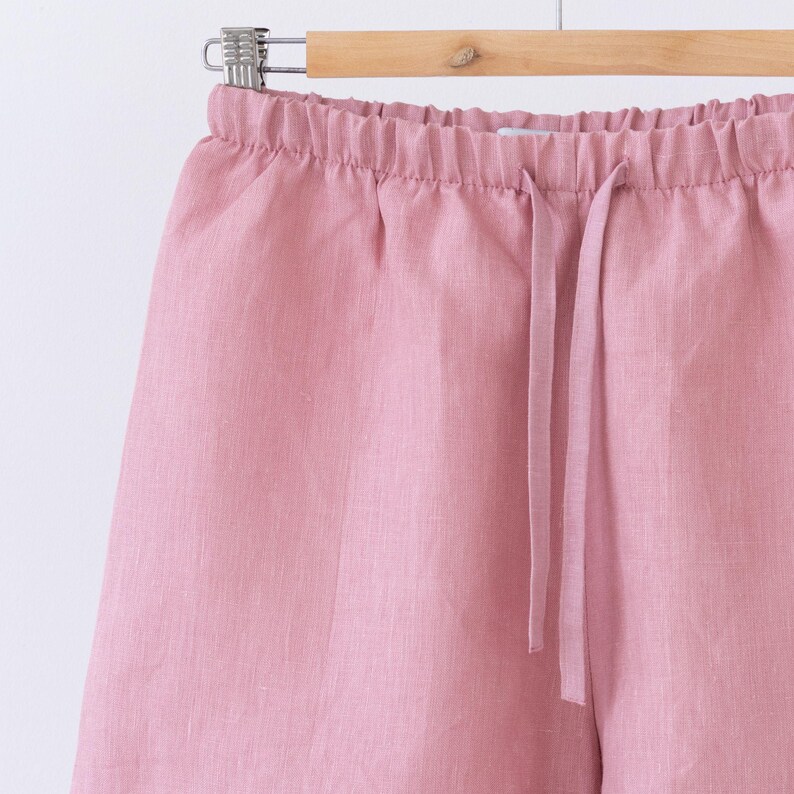 May include: Close-up of pink linen shorts hanging on a wooden hanger. The shorts have an elastic waistband with a drawstring. The fabric has a slightly textured appearance.