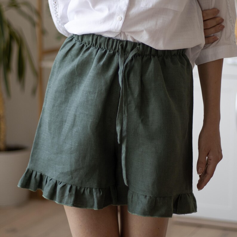 May include: Olive green linen shorts with a drawstring waist and ruffled hem. The shorts are made of a lightweight fabric and have a relaxed fit. The shorts are shown with a white button-down shirt.