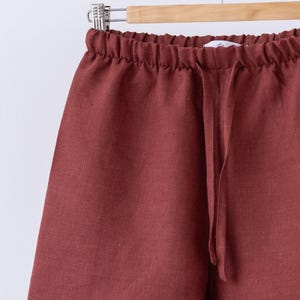 May include: Close-up of rust-colored linen shorts with an elastic waistband and drawstring. The shorts are hanging on a wooden hanger against a white background. The fabric has a textured appearance.