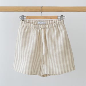 May include: Beige and white vertical striped shorts featuring an elasticated waistband with a matching fabric drawstring tie. The interior label displays the text BEANCHY and the size S. The garment is suspended from a wooden hanger.
