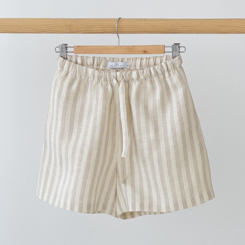 May include: Beige and white vertical striped shorts featuring an elasticated waistband with a matching fabric drawstring tie. The interior label displays the text BEANCHY and the size S. The garment is suspended from a wooden hanger.
