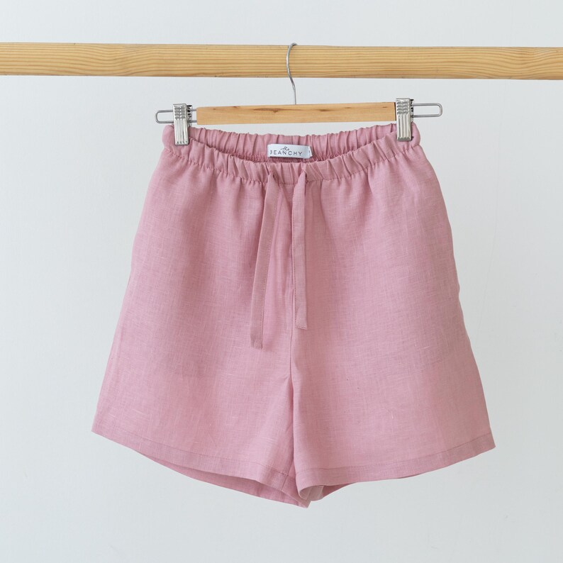 May include: Dusty rose shorts featuring an elasticated waistband with a matching drawstring tie. The garment includes a small white label at the centre of the inner waistband with the text "BEANCHY" printed in black, hanging from a wooden hanger.