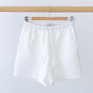 May include: Solid white shorts featuring an elasticated waistband with a matching fabric drawstring tie. The garment includes a small interior label with the text BEANCHY printed in black, hanging from a wooden hanger against a plain background.