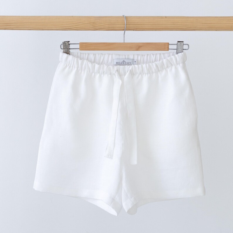 May include: Solid white shorts featuring an elasticated waistband with a matching fabric drawstring tie. The garment includes a small interior label with the text BEANCHY printed in black, hanging from a wooden hanger against a plain background.