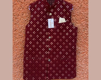 Op de afbeelding: Een bordeauxrode gilet met een geometrisch patroon van kleine gouden vierkantjes. De gilet heeft een pochet, een label en vier zilveren knopen. De gilet is mouwloos en heeft een donkere voering.