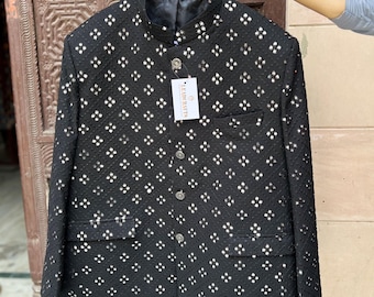 Op de afbeelding: Zwarte jas met een opstaande kraag en een patroon van kleine, reflecterende vierkantjes. De jas heeft een knoopsluiting, twee zakken en een label met de tekst "LEATHERUTS". De jas hangt aan een paarse hanger.