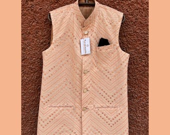 May include: A peach-colored, sleeveless vest with a mandarin collar and a geometric pattern. The vest has a pocket on the left side and is adorned with small, reflective embellishments. A tag with the text "PEARL KURTIS" is attached.