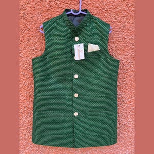 Op de afbeelding: Een mouwloos, smaragdgroen vest met een textuurpatroon en een opstaande kraag. Het vest heeft vier goudkleurige knopen, een kleine zak en een label. Het hangt aan een paarse hanger.