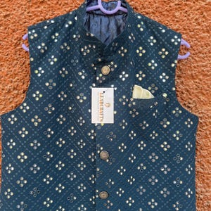 May include: A teal-colored, sleeveless vest with a repeating pattern of small, reflective embellishments. The vest has a mandarin collar, a pocket square, and decorative buttons. A tag reads "LEADCRAFTS".