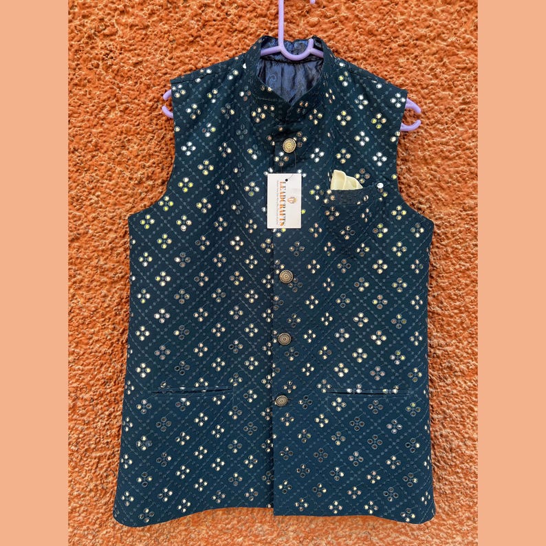 May include: A navy blue, sleeveless vest with a geometric pattern of small, reflective squares. The vest has a mandarin collar, a pocket, and gold-colored buttons. A white tag with black text is attached to the vest.