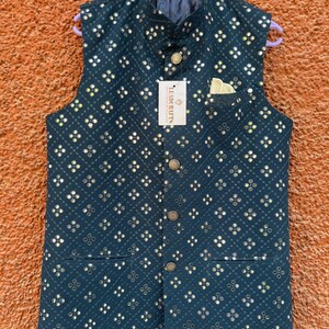 May include: A dark teal vest with a repeating pattern of small, reflective squares. The vest has a mandarin collar, a pocket square, and gold-colored buttons. A tag reads "LEADRAFTS". The vest is hanging on a purple hanger.