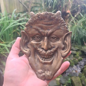 Può includere: Una placca da parete in ottone raffigurante un diavolo sorridente con corna, baffi e barba appuntita. Gli occhi del diavolo sono spalancati e la bocca è aperta in un ampio sorriso, mostrando i denti.