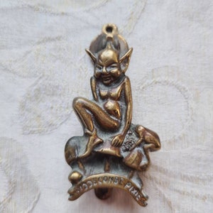 Könnte beinhalten: Ein Türklopfer aus Messing in Form eines Kobolds, der auf einem Pilz sitzt. Der Kobold hält einen kleinen Vogel in der Hand. Der Türklopfer trägt die Aufschrift "Widdecombe Pixie".