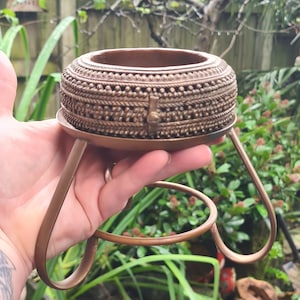 May include: A decorative brass stand with a round top and three legs. The top of the stand is intricately decorated with a repeating pattern of small circles and lines.