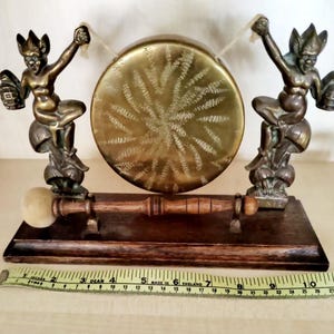 Puede incluir: Un gong de latón con dos figuras decorativas a cada lado. El gong está montado en un soporte de madera con un mazo de madera.