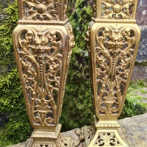 May include: A pair of ornate gold metal vases with intricate designs. The vases have a square base and a tall, slender body with a decorative top.