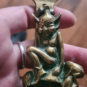 Puede incluir: Un llamador de puerta de latón con un diseño detallado de un duende sentado en un hongo. El llamador tiene un anillo para golpear y una pancarta con la inscripción "Haddecombe Pixie".