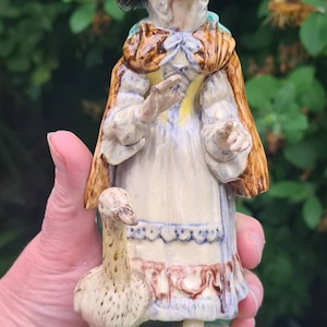 Può includere: Una statuetta in ceramica di una donna che indossa un cappello marrone e un vestito marrone e blu, che tiene un'oca. La statuetta è dipinta con colori marrone, blu, giallo e verde.